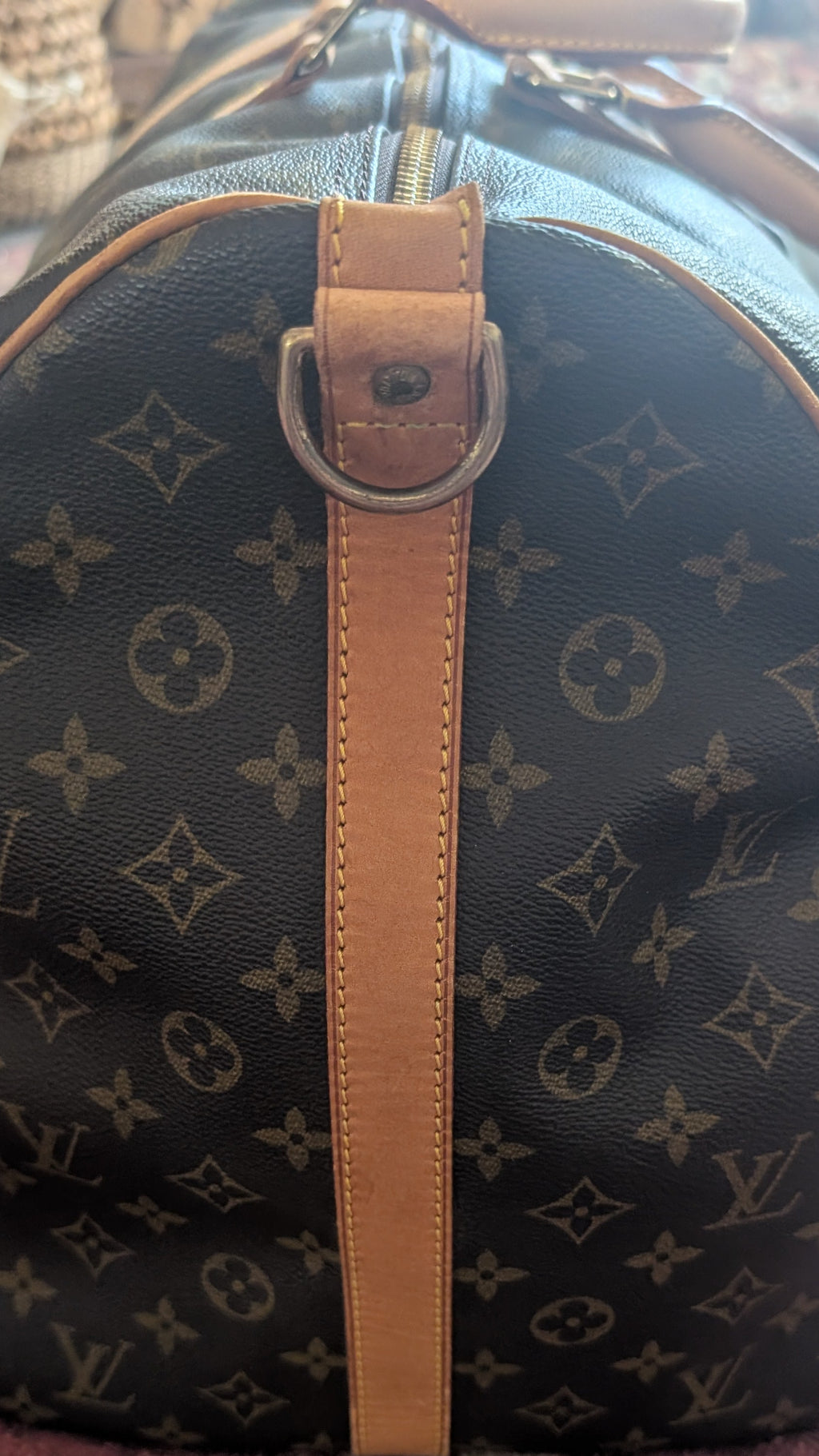 A Louis Vuitton monogram canvas duffel bag with tan leather trim and a central strap, photographed on a patterned rug. Pre-Owned Louis Vuitton Keepall Bandoulière 60 Monogram Travel Bag