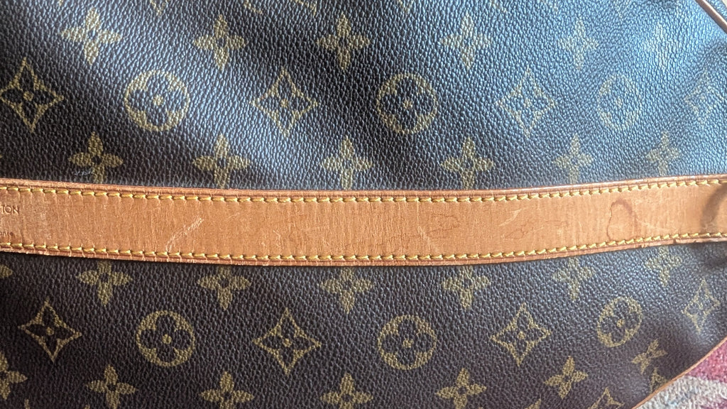 Close-up of a Louis Vuitton monogram canvas bag with a tan leather strap and metal hardware, resting on a patterned red and white surface. Pre-Owned Louis Vuitton Keepall Bandoulière 60 Monogram Travel Bag
