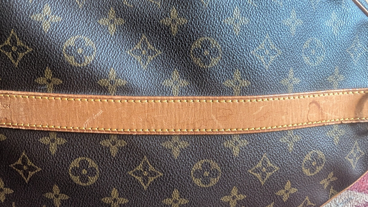 Close-up of a Louis Vuitton monogram canvas bag with a tan leather strap and metal hardware, resting on a patterned red and white surface. Pre-Owned Louis Vuitton Keepall Bandoulière 60 Monogram Travel Bag