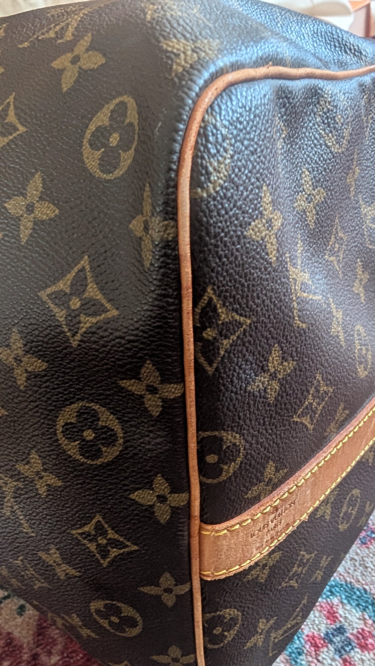 Close-up of a Louis Vuitton monogram canvas bag corner showing dark brown coated canvas with tan monogram motifs, tan leather piping, and a tan leather trim piece. Pre-Owned Louis Vuitton Keepall Bandoulière 60 Monogram Travel Bag