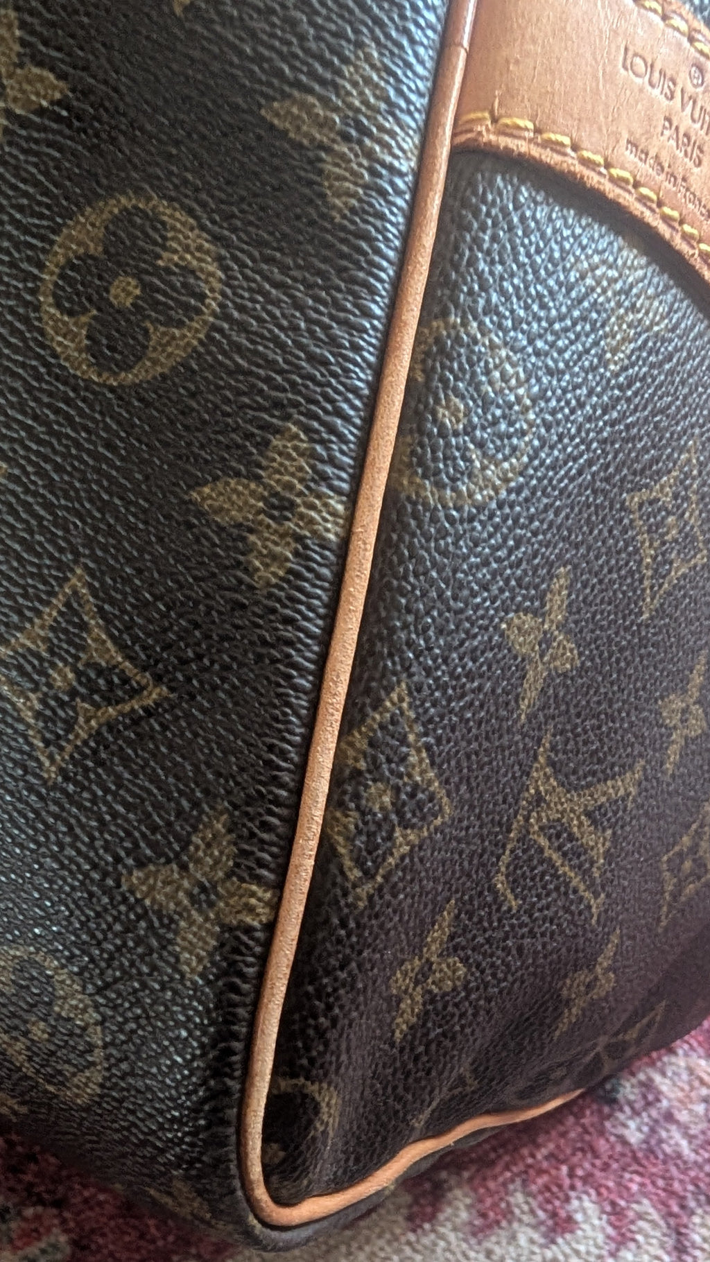 Close-up of a Louis Vuitton monogram canvas bag corner with tan leather trim, resting on a patterned rug. Pre-Owned Louis Vuitton Keepall Bandoulière 60 Monogram Travel Bag