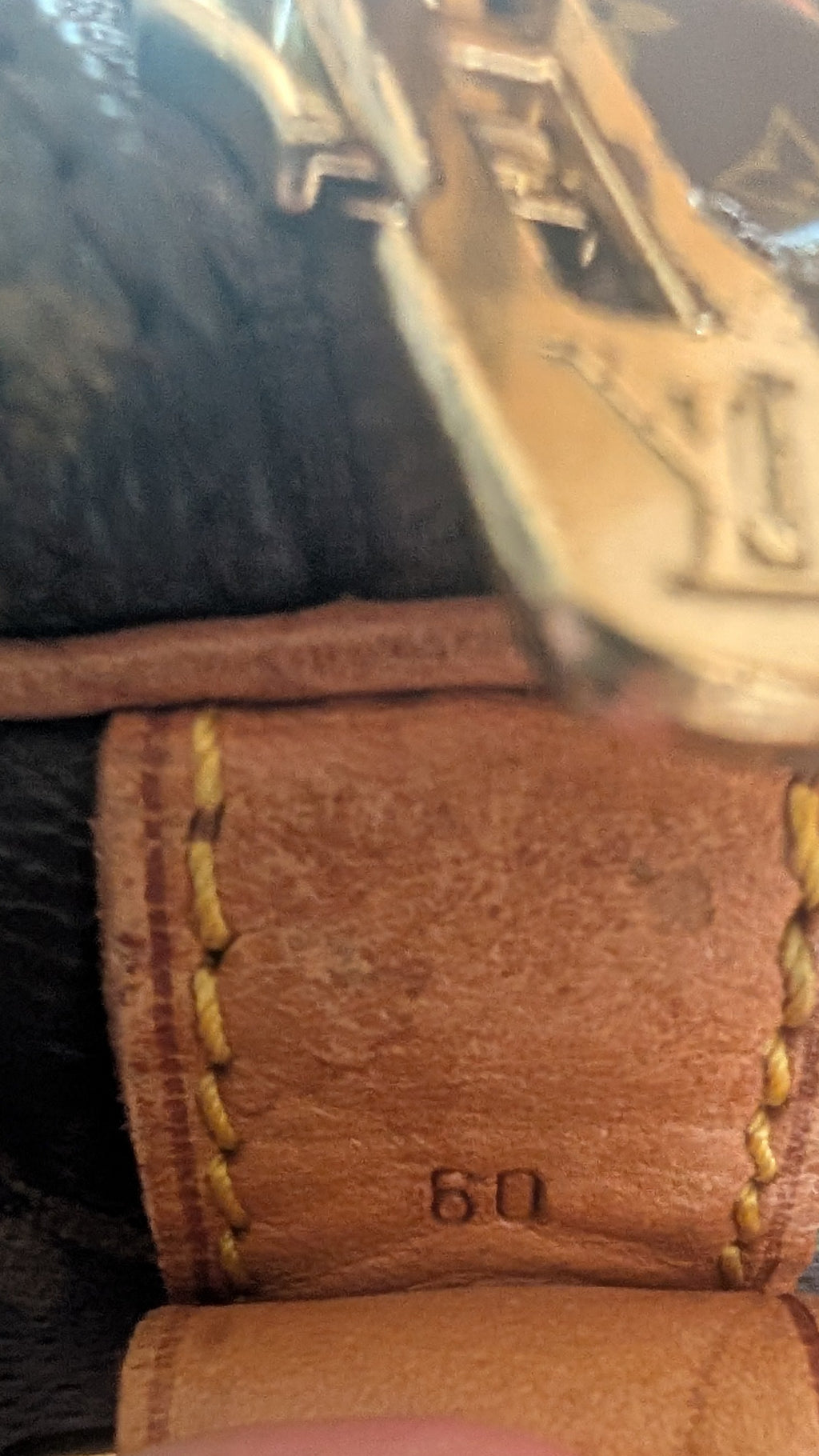 Close-up of a Louis Vuitton monogram bag strap with a finger pointing to the embossed size '60' on a tan leather tab, showing the brown monogram canvas and gold-tone hardware. Pre-Owned Louis Vuitton Keepall Bandoulière 60 Monogram Travel Bag