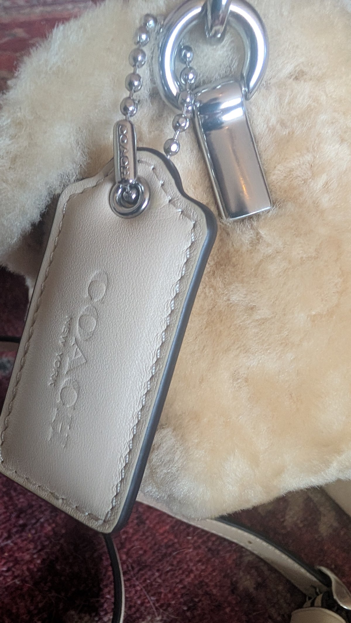 A Coach leather keychain tag in light beige with silver-tone hardware, shown close up on a cream-colored textured surface. Pre-Owned Coach Tabby 18 Shearling Shoulder / Crossbody Bag – Cream