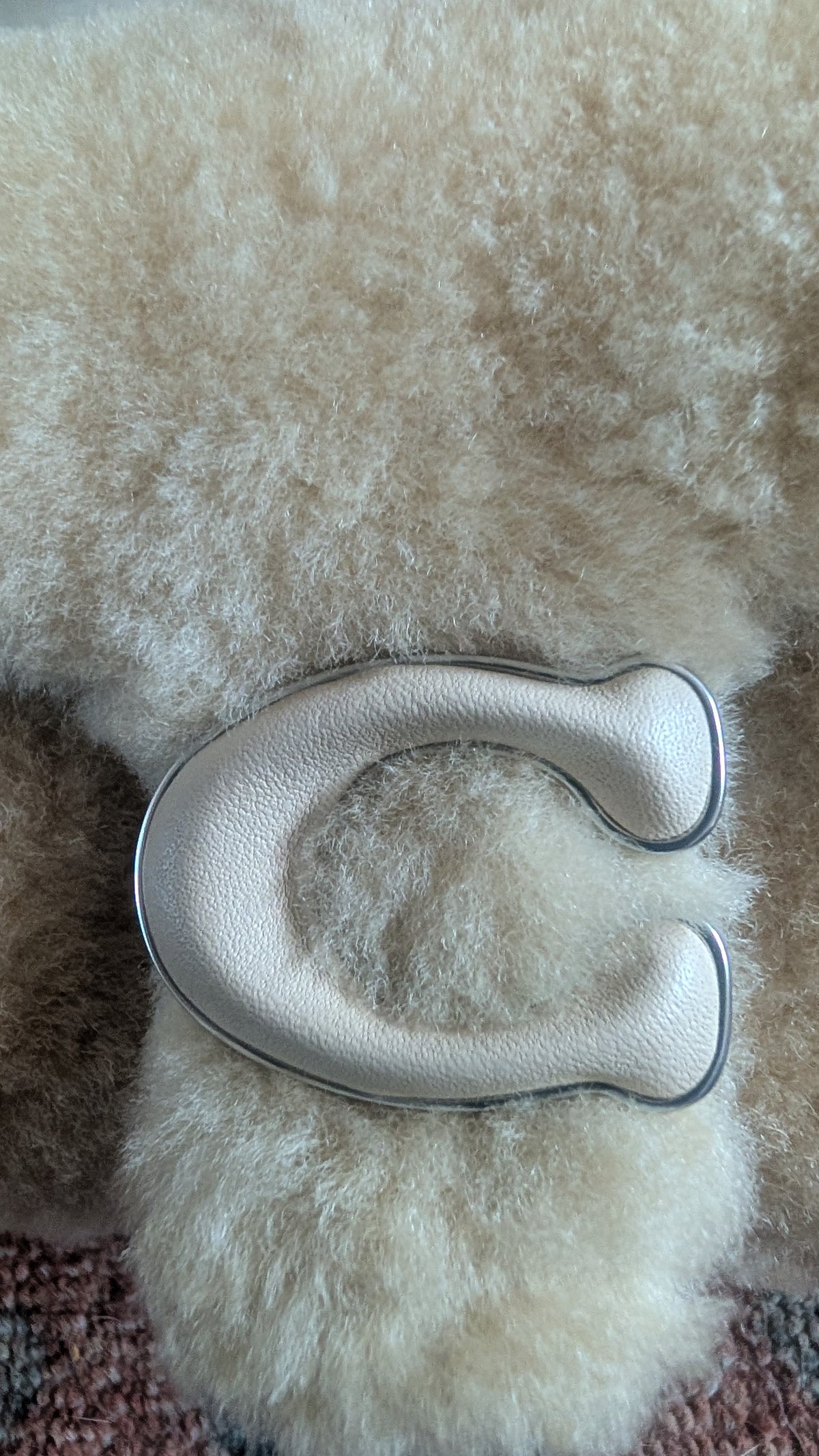 A close-up of a curved, light beige textured leather accessory with a silver metallic edge, resting on a fluffy cream sheepskin surface. Pre-Owned Coach Tabby 18 Shearling Shoulder / Crossbody Bag – Cream