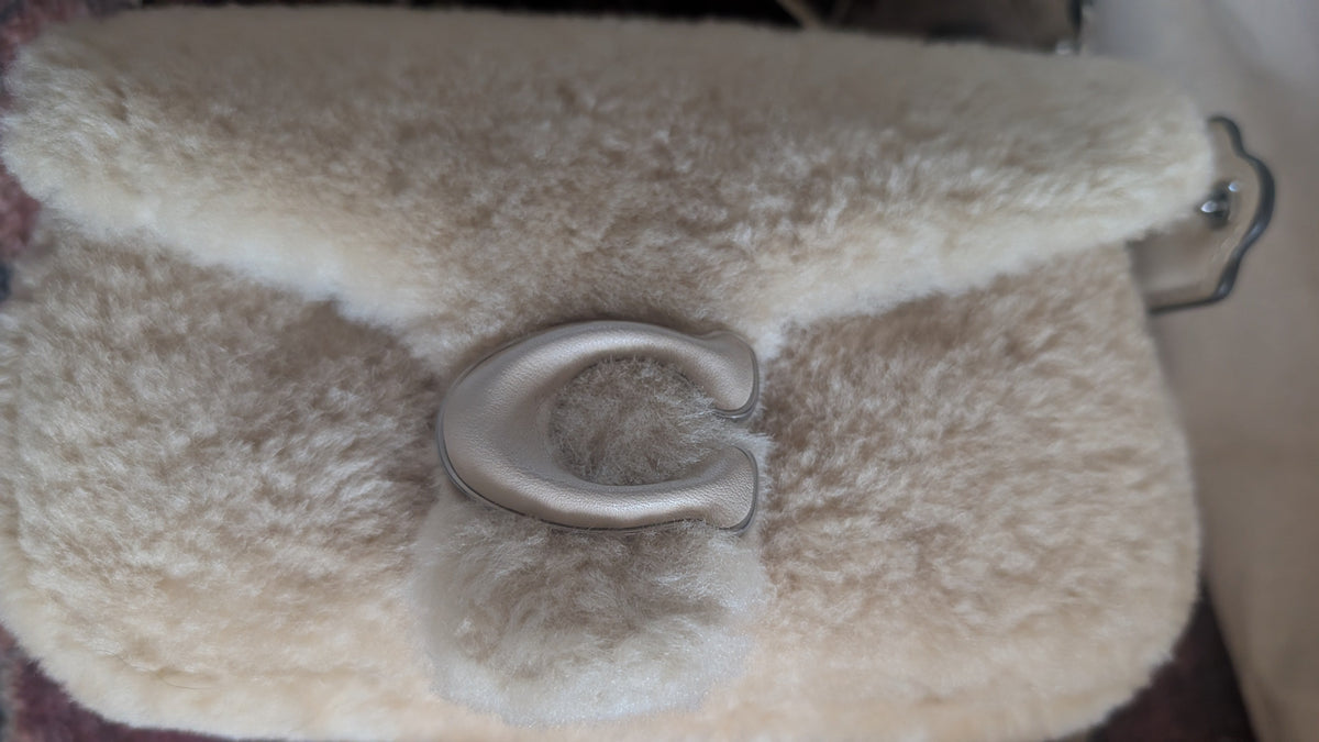 A close-up of a cream-colored sheepskin or faux-shearling handbag with a large silver metal horseshoe-shaped clasp. Pre-Owned Coach Tabby 18 Shearling Shoulder / Crossbody Bag – Cream