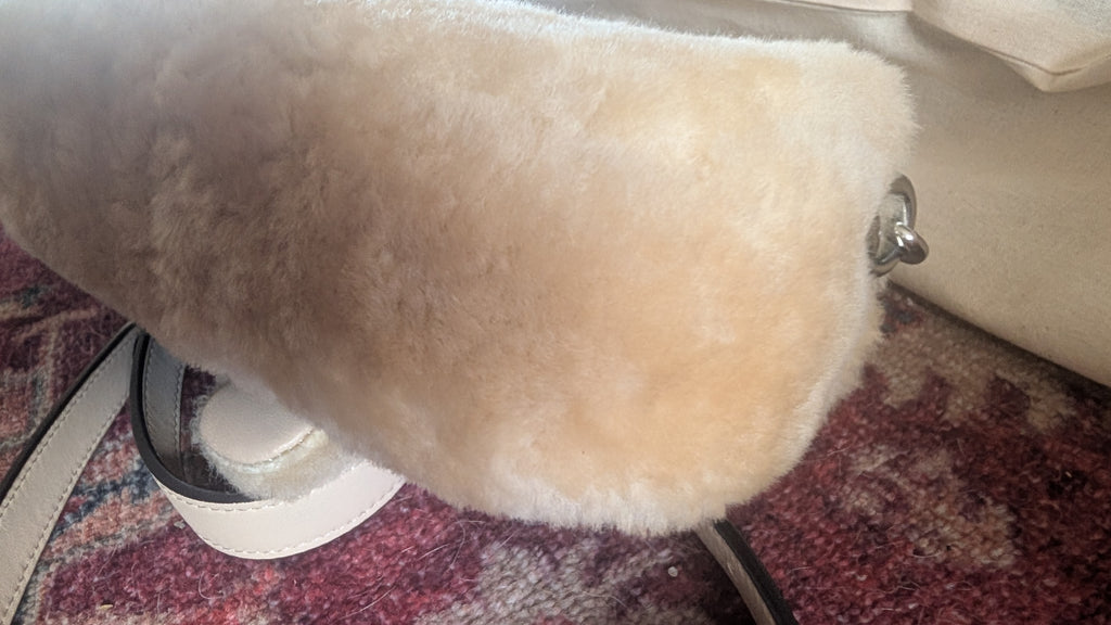 A close-up of a cream-colored sheepskin or faux-sheepskin bag or accessory, showing the soft textured material and a small silver metal stud detail, resting on a patterned rug. Pre-Owned Coach Tabby 18 Shearling Shoulder / Crossbody Bag – Cream