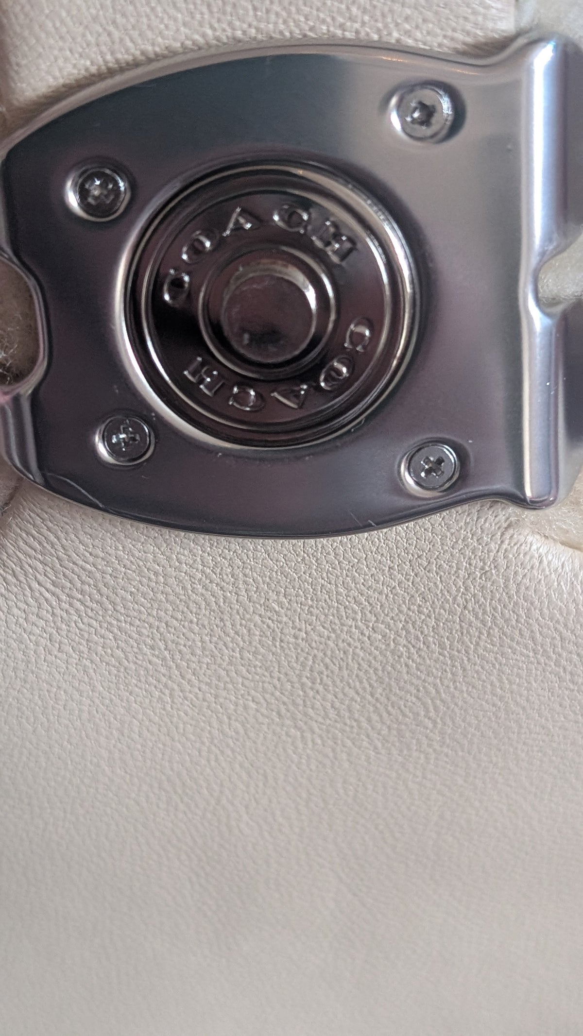 Close-up of a Coach metal buckle closure on a light beige textured leather surface, with the Coach logo embossed in the center. Pre-Owned Coach Tabby 18 Shearling Shoulder / Crossbody Bag – Cream
