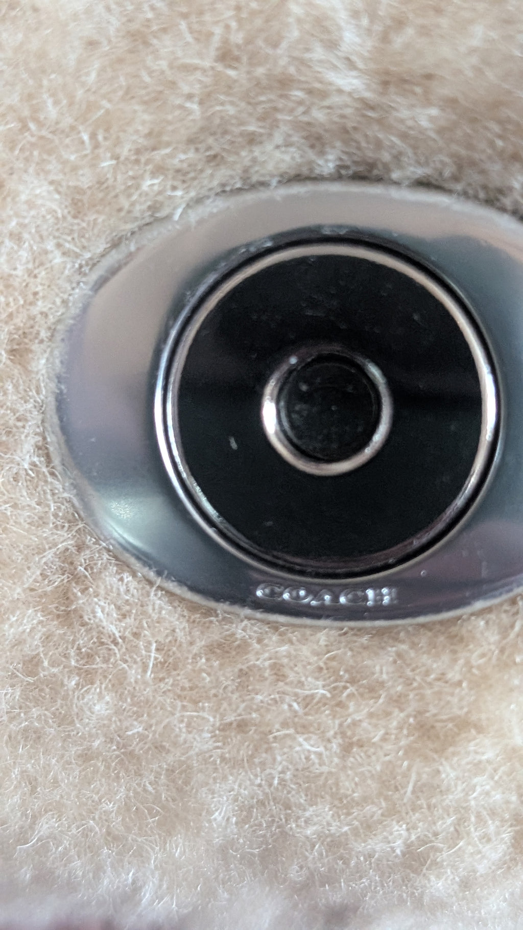 Close-up of a Coach oval metal emblem with a black center and silver border, set against a beige textured faux-fur background. Pre-Owned Coach Tabby 18 Shearling Shoulder / Crossbody Bag – Cream