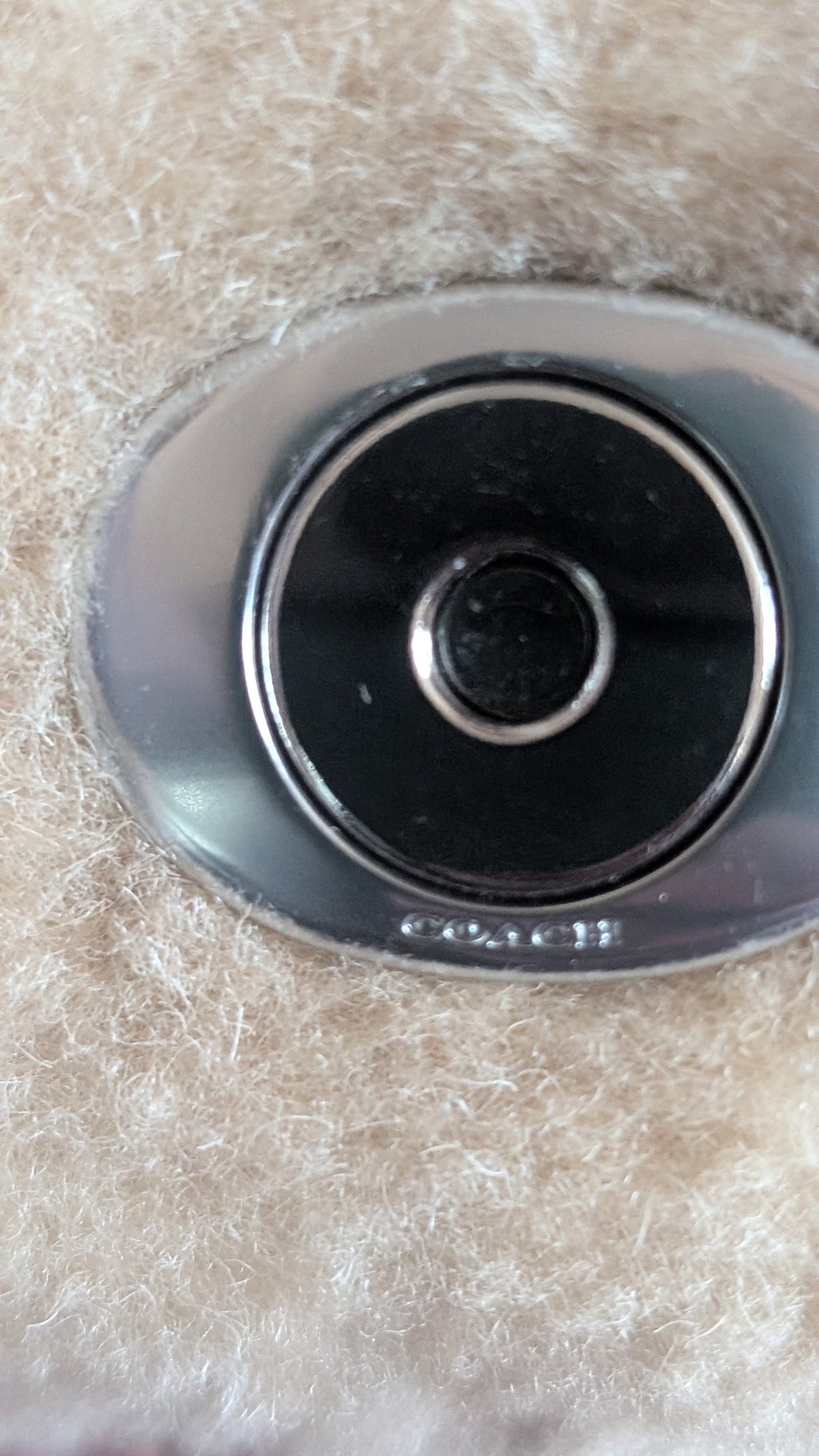 Close-up of a Coach oval metal emblem with a black center and silver border, set against a beige textured faux-fur background. Pre-Owned Coach Tabby 18 Shearling Shoulder / Crossbody Bag – Cream