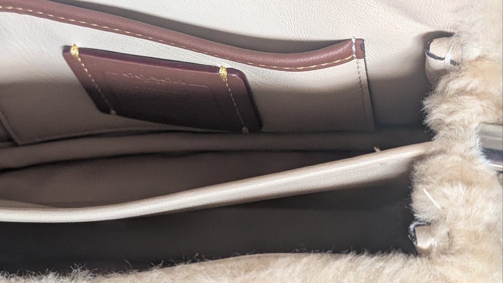 Close-up of the interior of a handbag showing a cream-colored leather lining, a brown leather pocket with gold-tone hardware, and a fluffy cream faux-fur trim. Pre-Owned Coach Tabby 18 Shearling Shoulder / Crossbody Bag – Cream