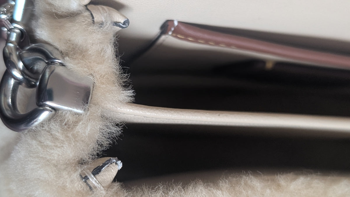 Close-up detail of a handbag showing a fluffy cream sheepskin or faux-shearling section attached to a silver-tone metal ring and a light beige strap, with a dark interior background. Pre-Owned Coach Tabby 18 Shearling Shoulder / Crossbody Bag – Cream