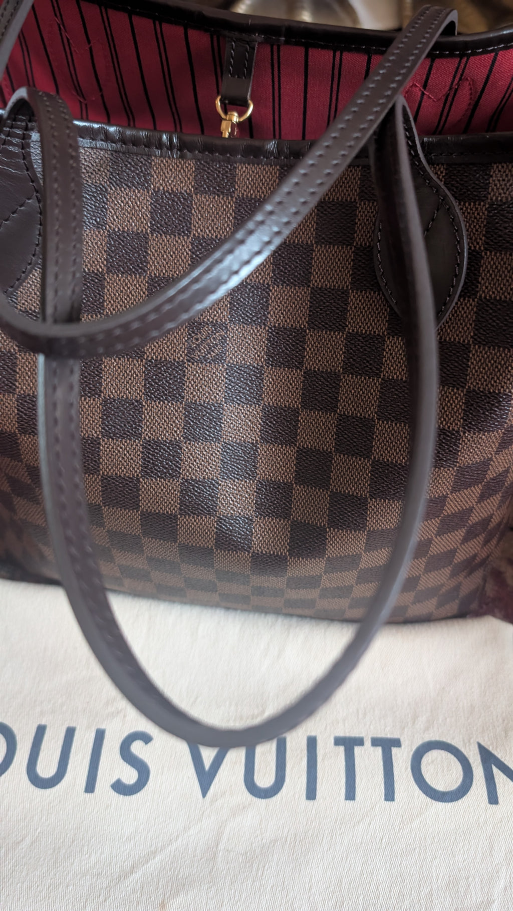A Louis Vuitton handbag in the Damier Ebene checkered pattern with dark brown leather handles, resting on a white dust bag with the Louis Vuitton logo partially visible. Pre-Owned Louis Vuitton Neverfull MM Damier Ebene, Cerise Red