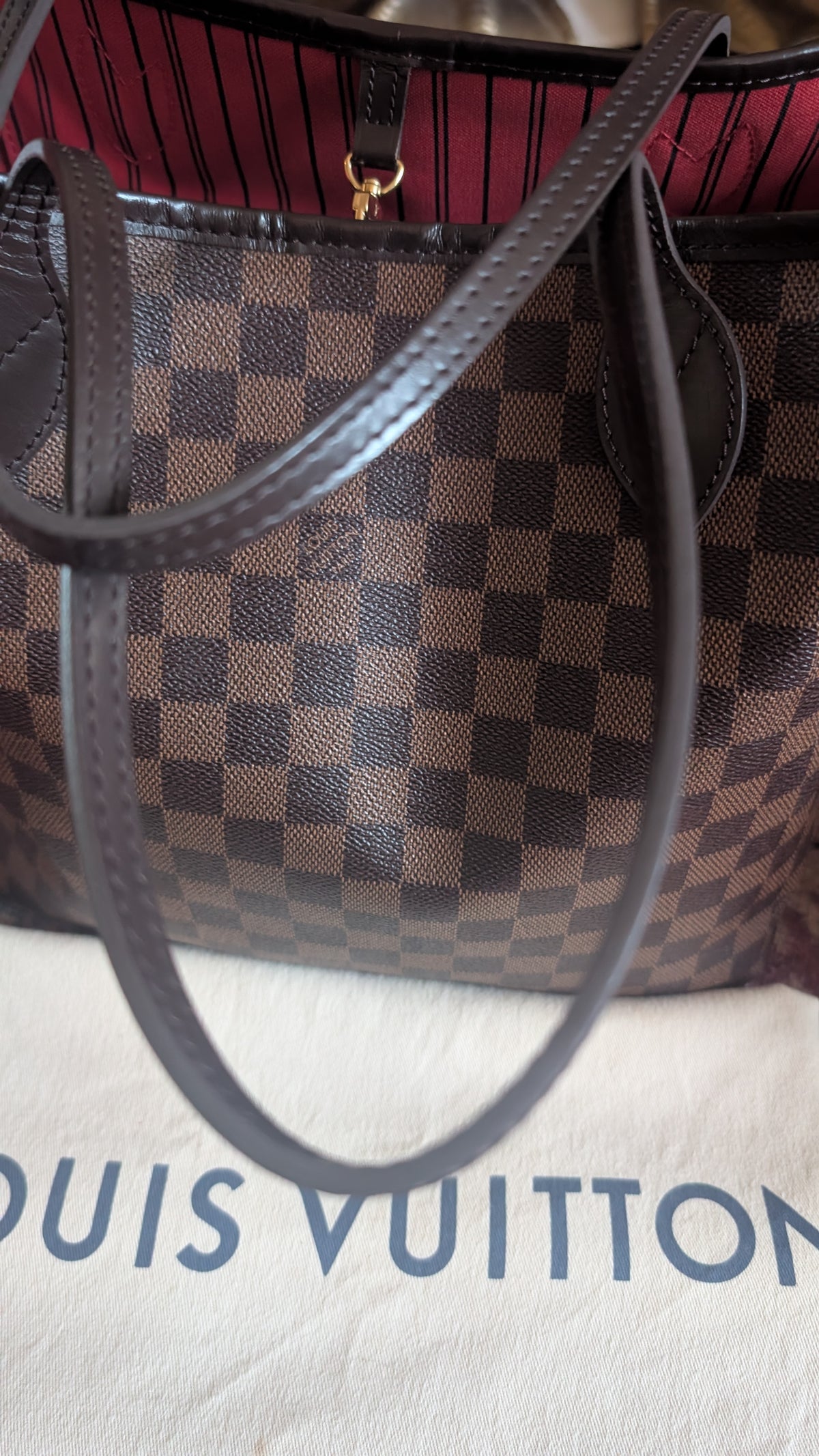 A Louis Vuitton handbag in the Damier Ebene checkered pattern with dark brown leather handles, resting on a white dust bag with the Louis Vuitton logo partially visible. Pre-Owned Louis Vuitton Neverfull MM Damier Ebene, Cerise Red