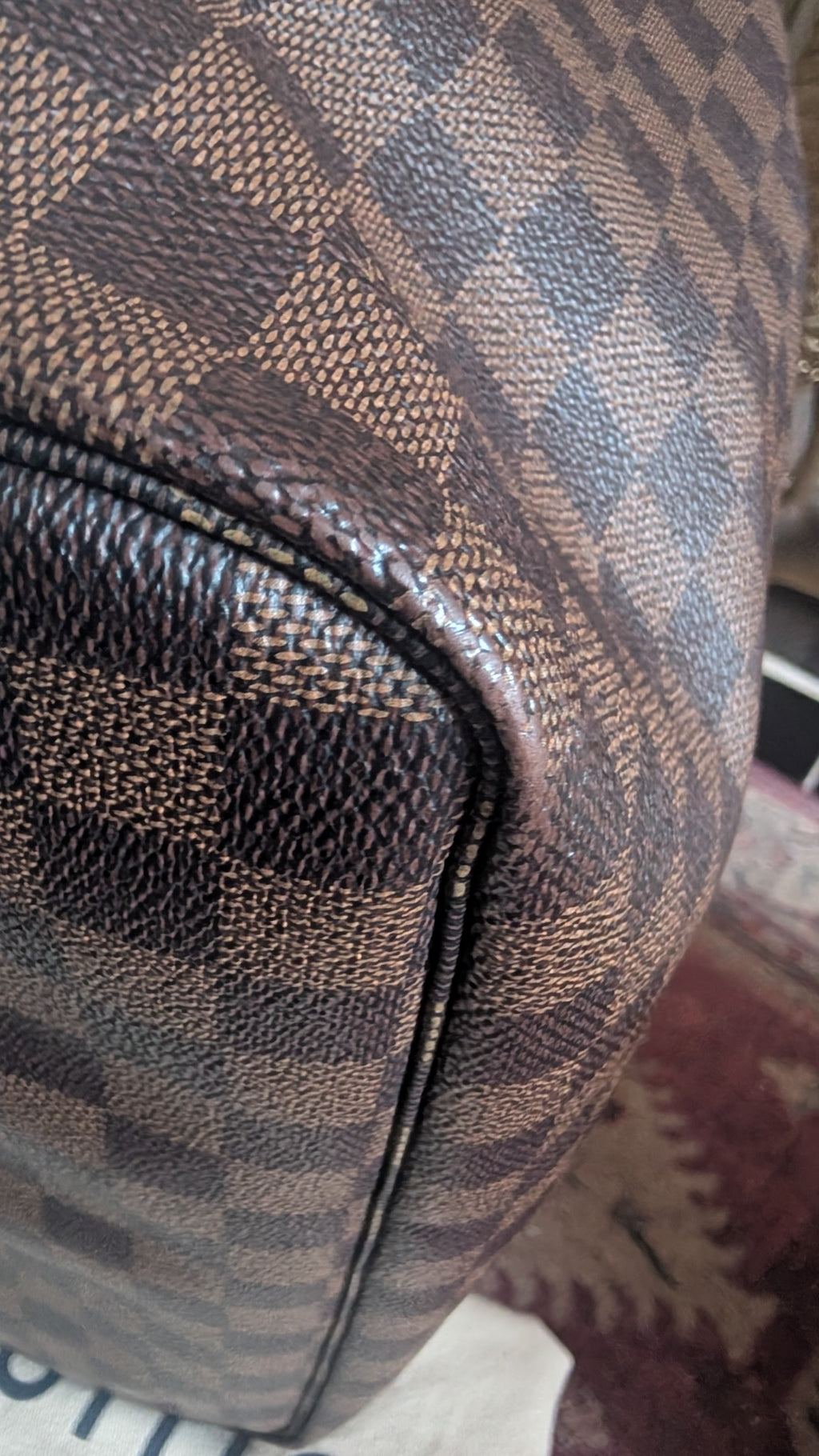 Close-up of a brown and beige checkered leather bag corner with pebbled texture and a black zipper. Pre-Owned Louis Vuitton Neverfull MM Damier Ebene, Cerise Red