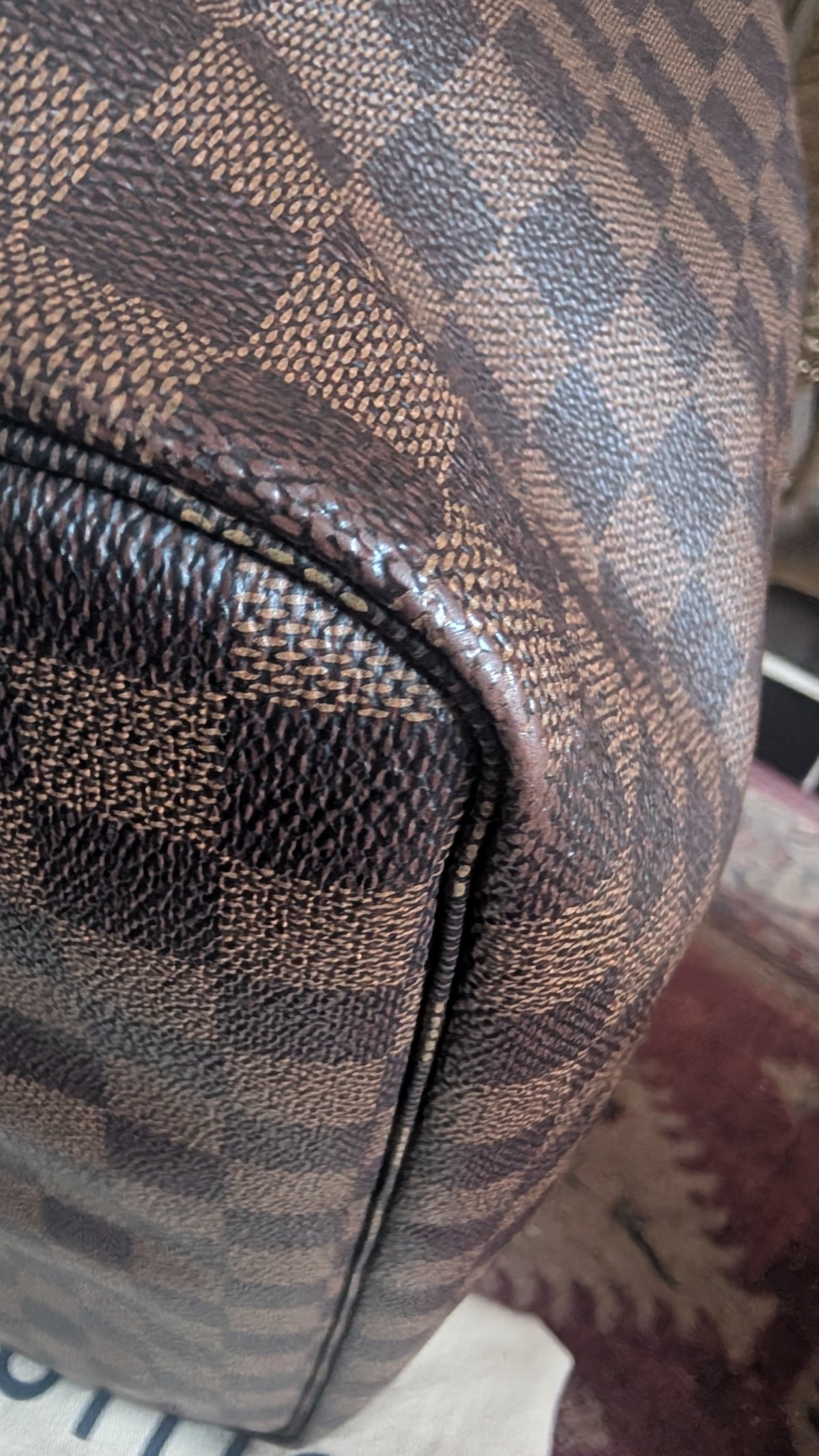 Close-up of a brown and beige checkered leather bag corner with pebbled texture and a black zipper. Pre-Owned Louis Vuitton Neverfull MM Damier Ebene, Cerise Red