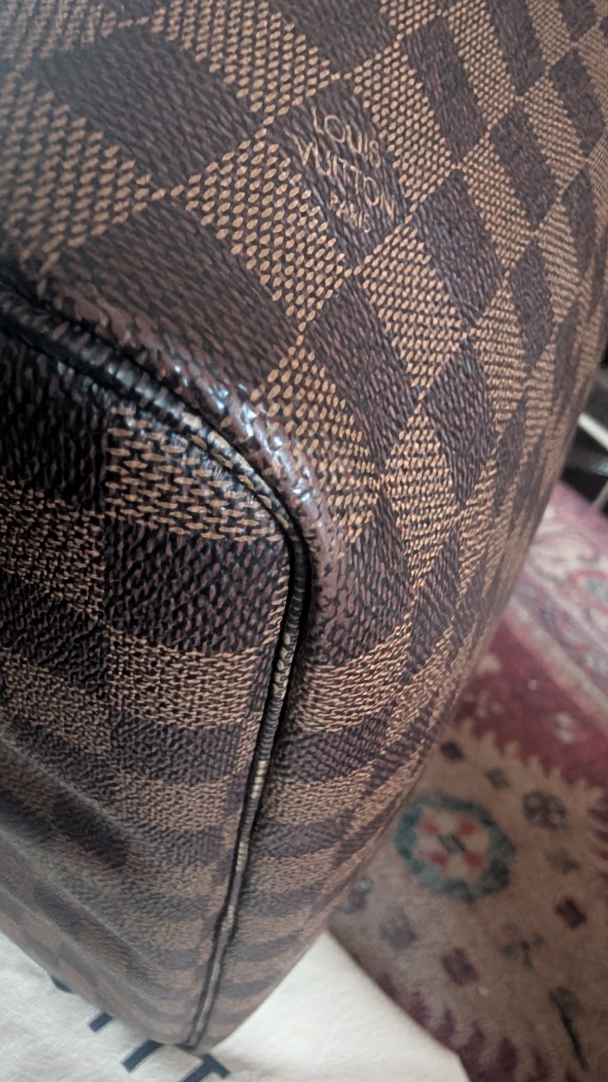 Close-up of a Louis Vuitton bag corner showing the brand's signature Damier Ebene checkered pattern in brown and beige, with a leather trim and a patterned rug in the background. Pre-Owned Louis Vuitton Neverfull MM Damier Ebene, Cerise Red