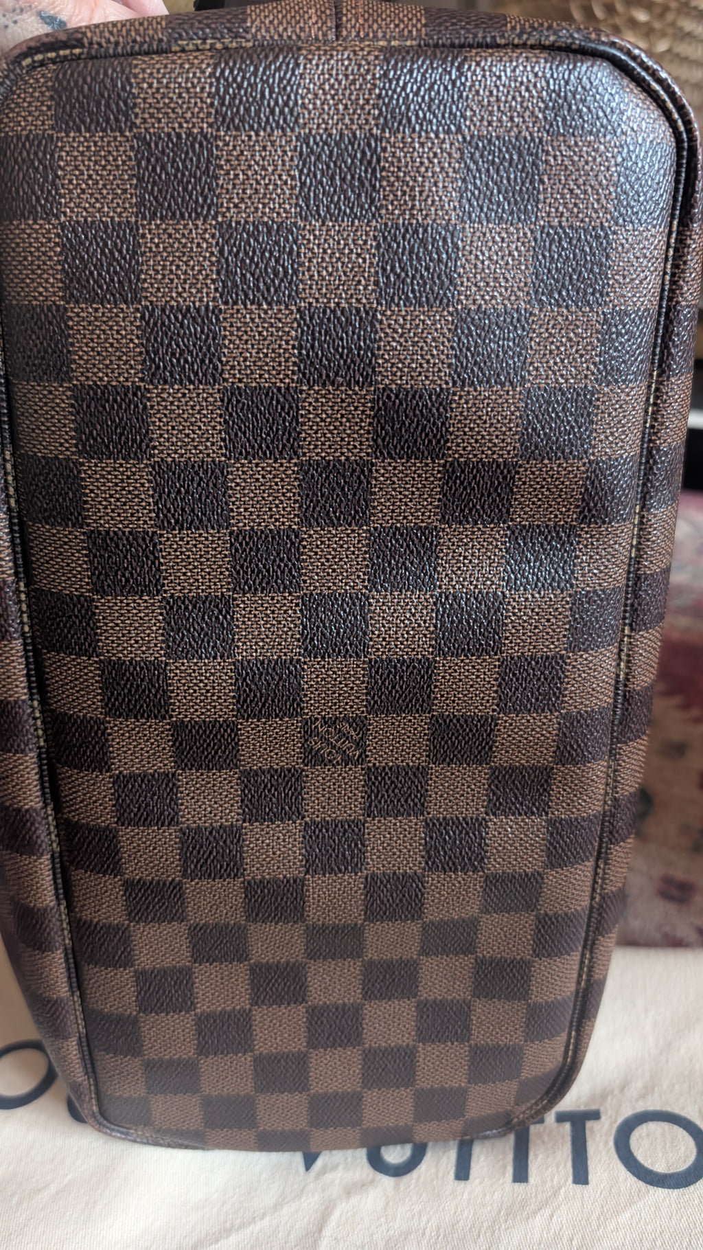 A close-up of a brown and dark brown checkerboard patterned leather bag, showing the textured Damier Ebene design. The Louis Vuitton brand name is partially visible on the white surface underneath. Pre-Owned Louis Vuitton Neverfull MM Damier Ebene, Cerise Red