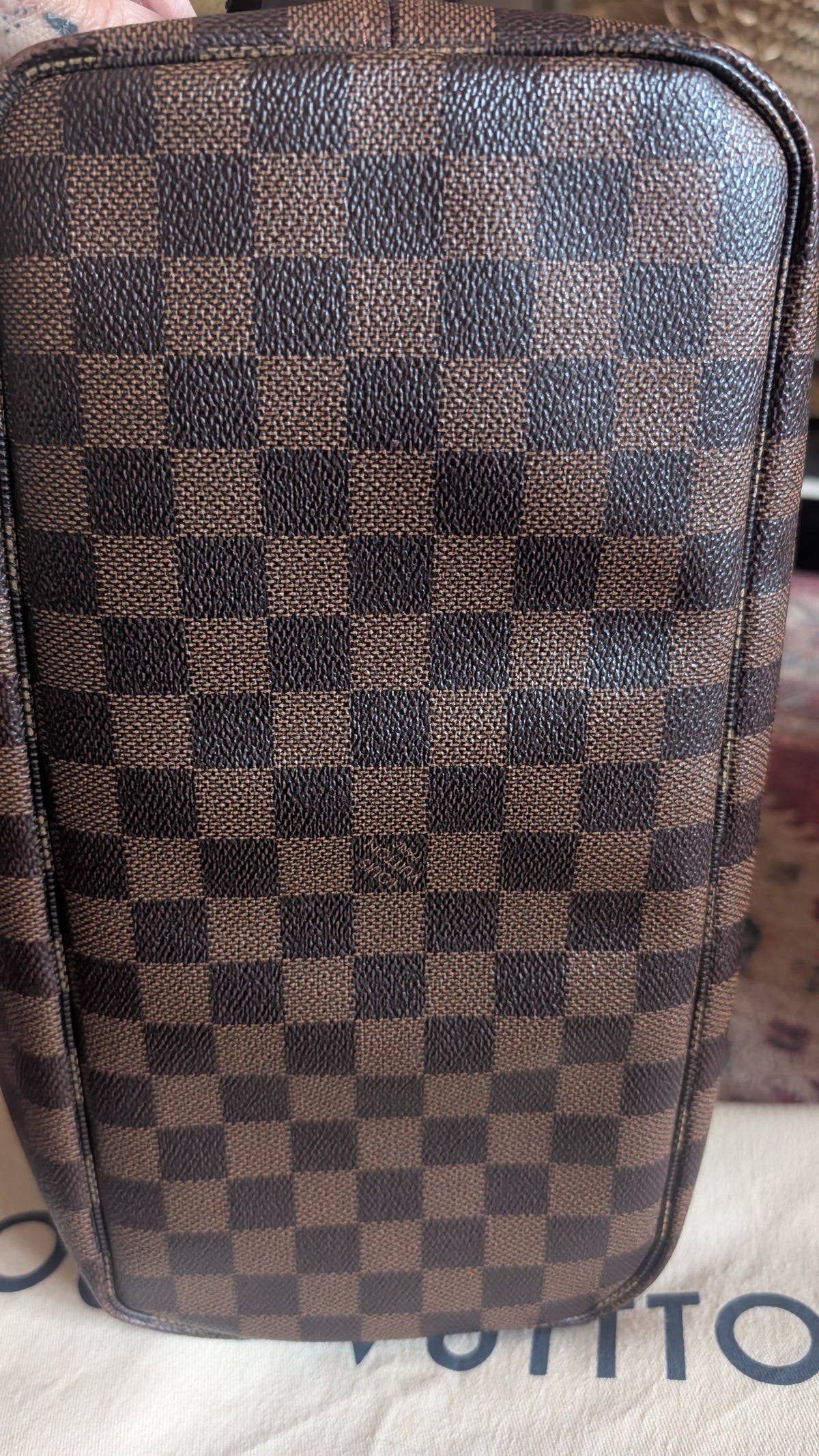 A close-up of a brown and dark brown checkerboard patterned leather bag, showing the textured Damier Ebene design. The Louis Vuitton brand name is partially visible on the white surface underneath. Pre-Owned Louis Vuitton Neverfull MM Damier Ebene, Cerise Red