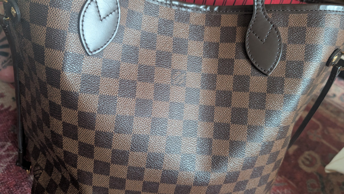Close-up of a brown and beige checkered canvas handbag with dark brown leather trim and leaf-shaped accents, photographed indoors on a patterned rug. Pre-Owned Louis Vuitton Neverfull MM Damier Ebene, Cerise Red