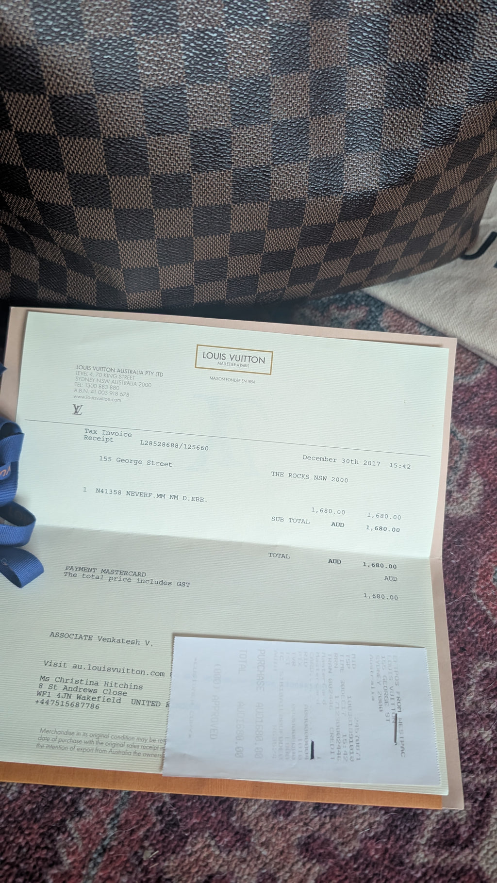 An open Louis Vuitton receipt or document folder placed on a patterned carpet, with a Louis Vuitton monogram bag visible in the background. Pre-Owned Louis Vuitton Neverfull MM Damier Ebene, Cerise Red