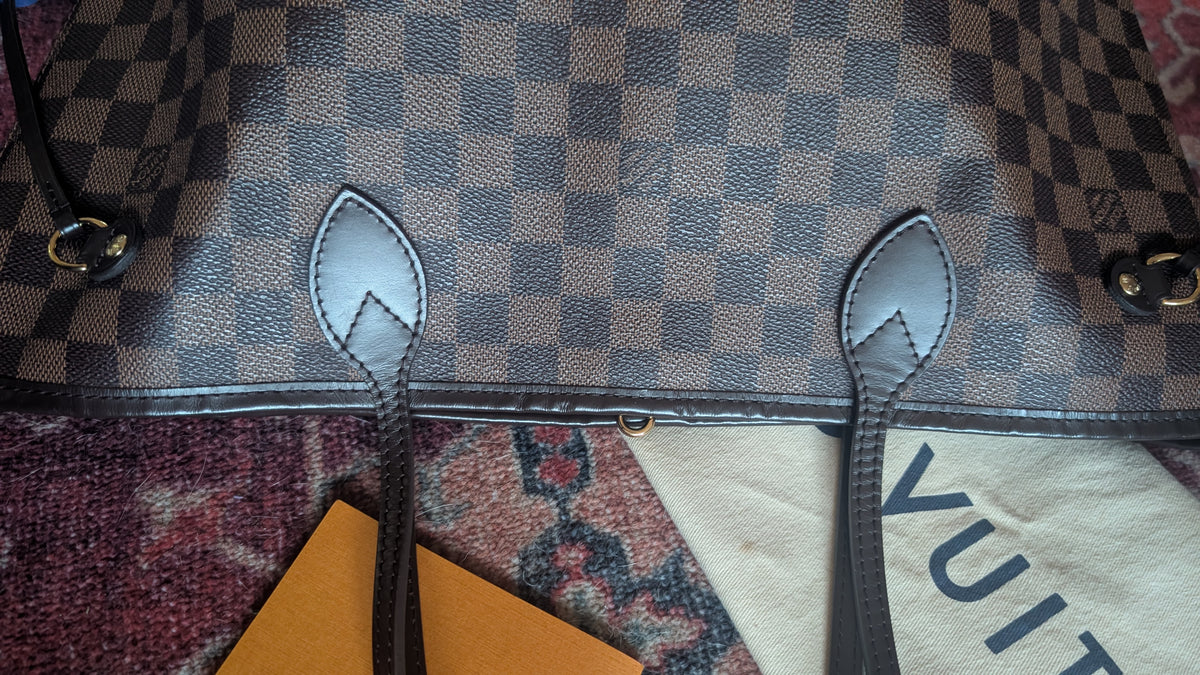 Close-up of a Louis Vuitton tote bag in Damier Ebene canvas, showing the brown and beige checkered pattern, dark brown leather trim, and hanging strap tabs. A dust bag and orange card are partially visible underneath on a patterned rug. Pre-Owned Louis Vuitton Neverfull MM Damier Ebene, Cerise Red