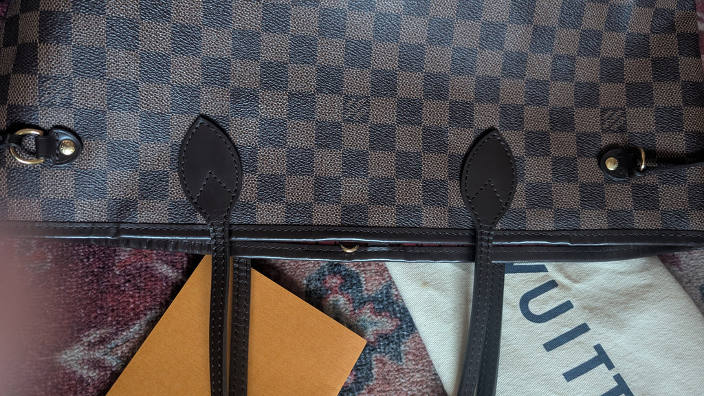Close-up of a Louis Vuitton handbag in Damier Ebene canvas, showing the dark brown and gray checkered pattern, black leather trim, and part of the white branded dust bag. Pre-Owned Louis Vuitton Neverfull MM Damier Ebene, Cerise Red