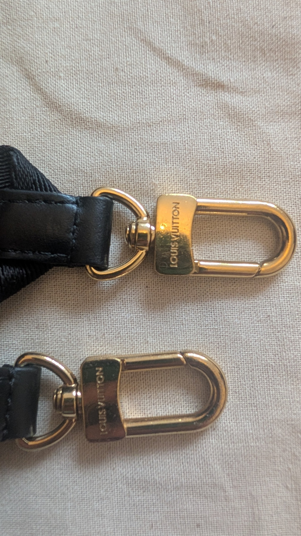 Close-up of a Louis Vuitton shoulder strap with black leather and gold-tone hardware, showing the engraved brand name on the clasp against a light fabric background. Pre-Owned Louis Vuitton Petite Malle Souple Monogram Bag