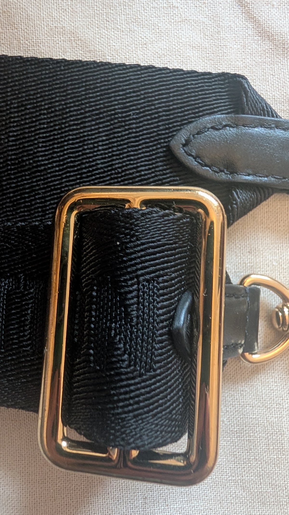 Close-up of a black woven strap with a large polished gold-tone rectangular buckle and a small gold-tone ring, laid on a light beige fabric surface. Pre-Owned Louis Vuitton Petite Malle Souple Monogram Bag