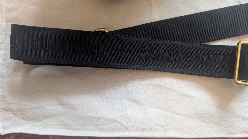 A black Louis Vuitton canvas shoulder strap with gold-tone hardware, laid flat on a white textured fabric background. Pre-Owned Louis Vuitton Petite Malle Souple Monogram Bag