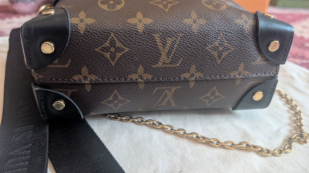 A Louis Vuitton monogram canvas shoulder bag with black leather trim and a gold-tone chain strap, resting on a white textured fabric surface. Pre-Owned Louis Vuitton Petite Malle Souple Monogram Bag