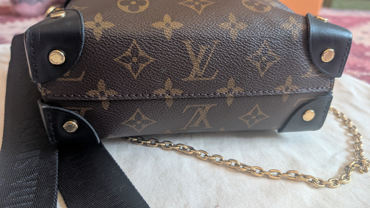 A Louis Vuitton monogram canvas shoulder bag with black leather trim and a gold-tone chain strap, resting on a white textured fabric surface. Pre-Owned Louis Vuitton Petite Malle Souple Monogram Bag