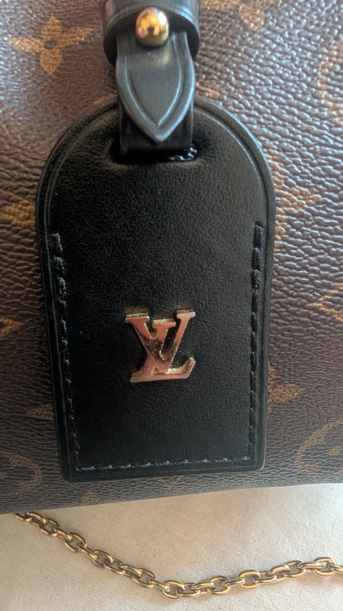 Close-up of a Louis Vuitton black leather luggage tag with a gold-tone LV logo, attached to a brown monogram bag and a gold chain. Pre-Owned Louis Vuitton Petite Malle Souple Monogram Bag