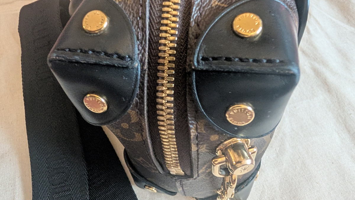 Close-up of a Louis Vuitton shoulder bag in brown monogram canvas with black leather trim, gold-tone zippers, and hardware, resting on a light fabric surface. Pre-Owned Louis Vuitton Petite Malle Souple Monogram Bag