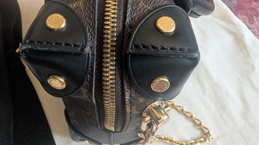 Close-up of a black leather and monogram canvas handbag with gold-tone hardware, including snap buttons, a central zipper, and a chain strap, resting on a light fabric surface. Pre-Owned Louis Vuitton Petite Malle Souple Monogram Bag