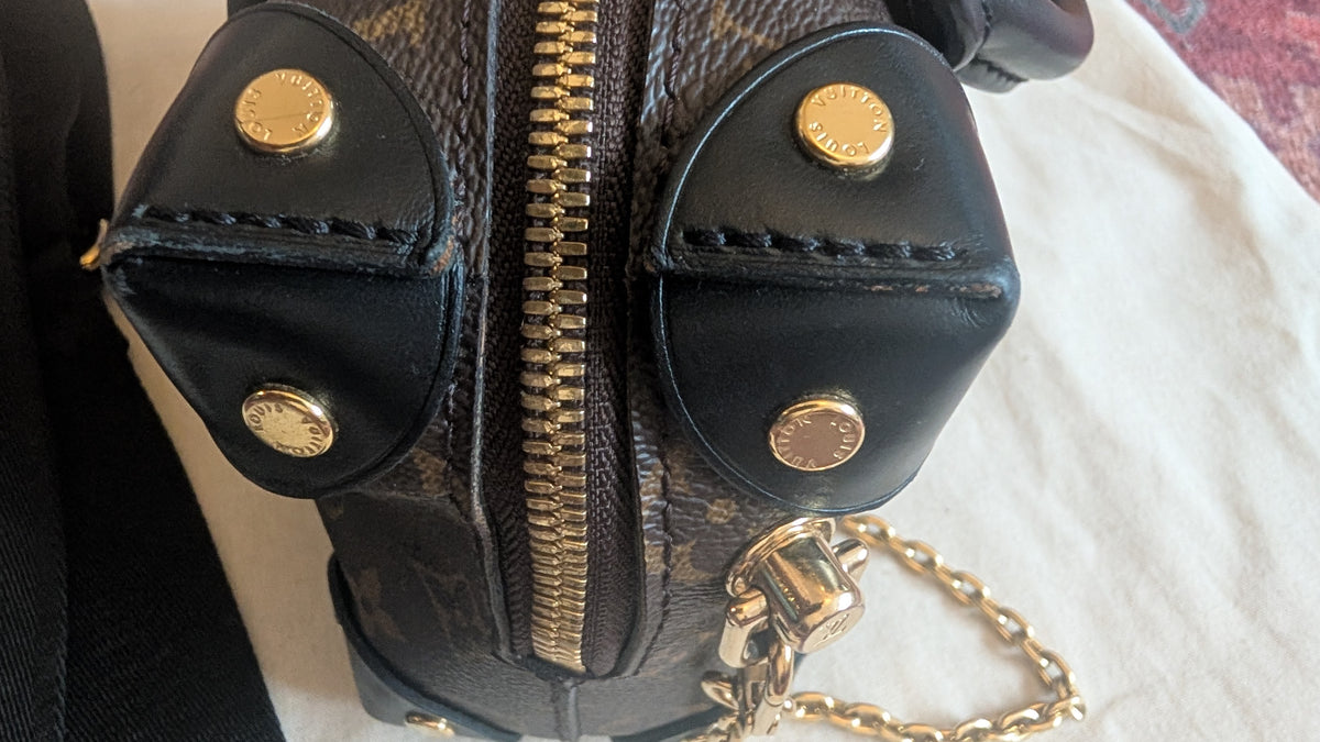 Close-up of a black leather and monogram canvas handbag with gold-tone hardware, including snap buttons, a central zipper, and a chain strap, resting on a light fabric surface. Pre-Owned Louis Vuitton Petite Malle Souple Monogram Bag