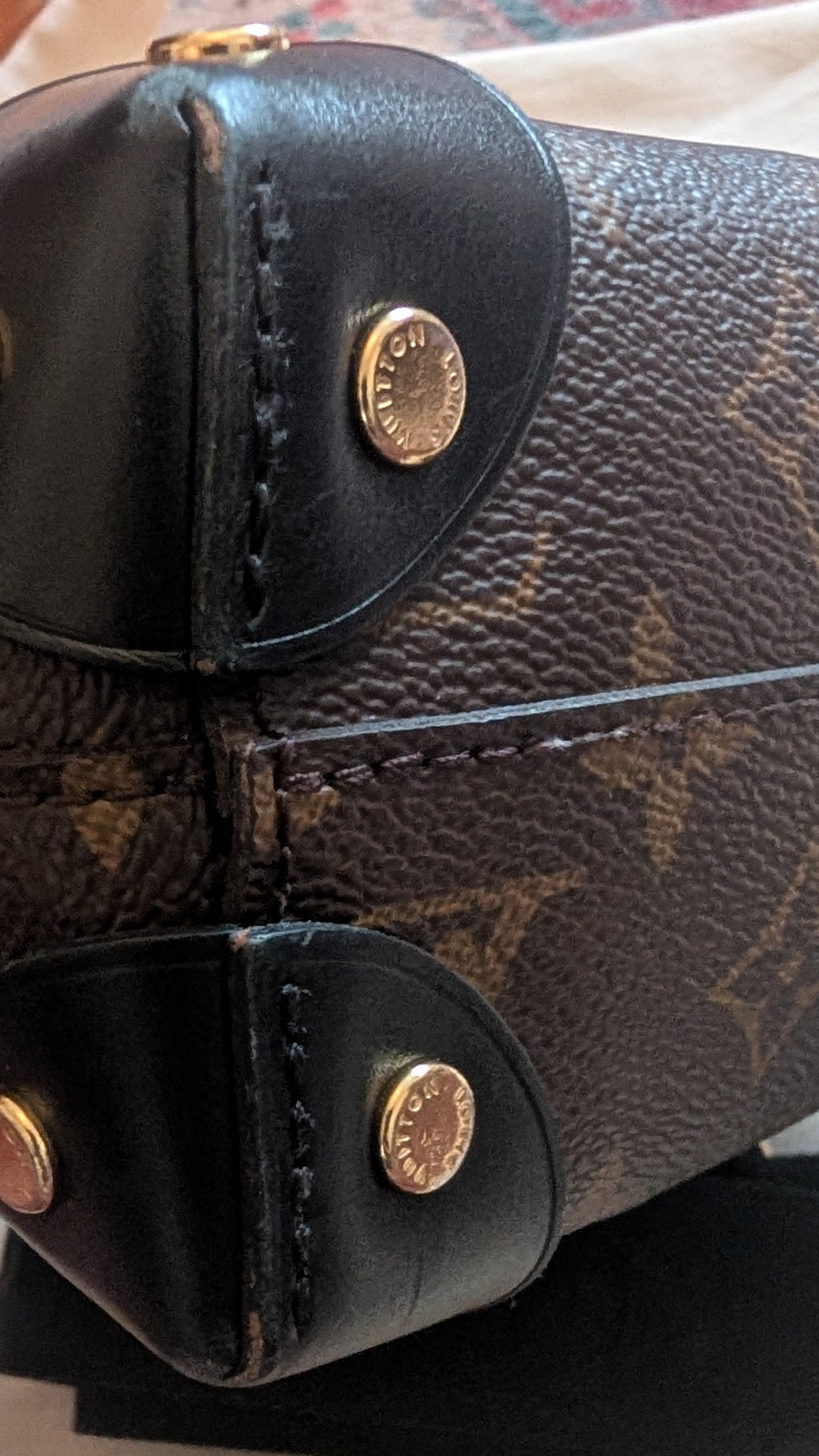 Close-up of a Louis Vuitton monogram handbag showing the bottom corner with black leather trim and gold-tone hardware, resting on a light-colored surface. Pre-Owned Louis Vuitton Petite Malle Souple Monogram Bag