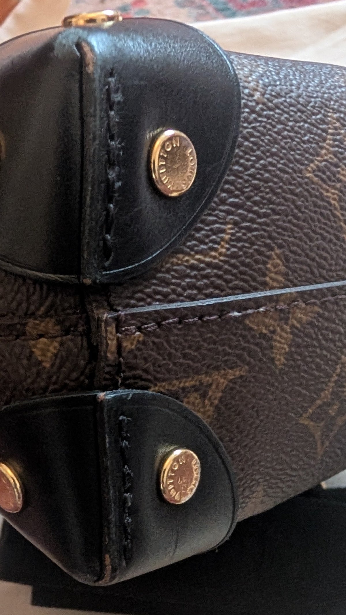 Close-up of a Louis Vuitton monogram handbag showing the bottom corner with black leather trim and gold-tone hardware, resting on a light-colored surface. Pre-Owned Louis Vuitton Petite Malle Souple Monogram Bag