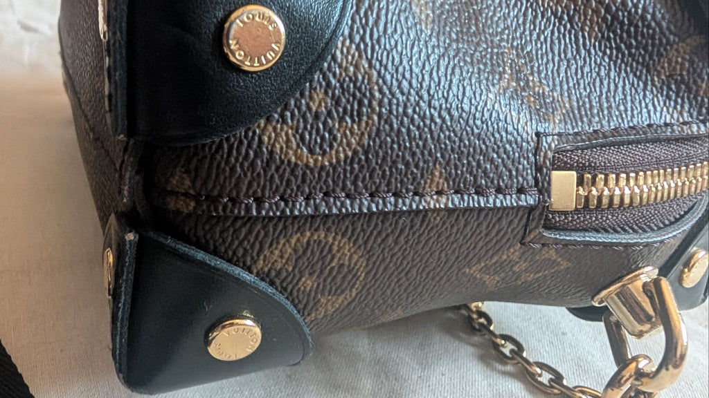 Close-up of a dark brown monogram handbag with gold-tone hardware, showing the textured coated canvas, leather trim, zipper, and chain strap on a light fabric surface. Pre-Owned Louis Vuitton Petite Malle Souple Monogram Bag