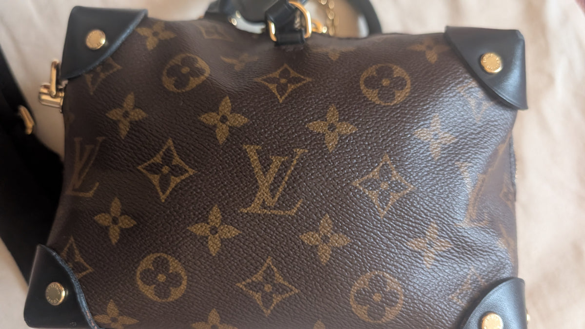 A close-up of a Louis Vuitton monogram canvas handbag in dark brown with black leather corner protectors and gold-tone hardware, resting on a light beige fabric surface. Pre-Owned Louis Vuitton Petite Malle Souple Monogram Bag
