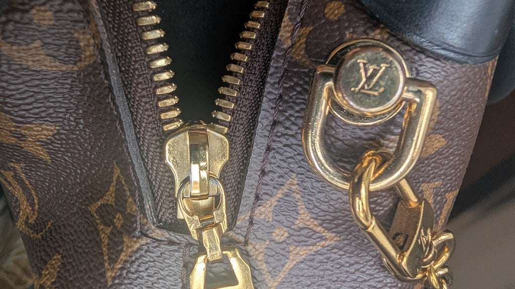 Close-up of a Louis Vuitton monogram handbag showing the brown coated canvas with gold-tone monogram print, a gold zipper pull, and a gold-tone metal ring with an LV logo. Pre-Owned Louis Vuitton Petite Malle Souple Monogram Bag