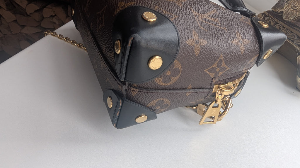 Close-up of a Louis Vuitton monogram canvas handbag corner with black leather trim, gold-tone hardware, and a chain strap on a white surface. Pre-Owned Louis Vuitton Petite Malle Souple Monogram Bag