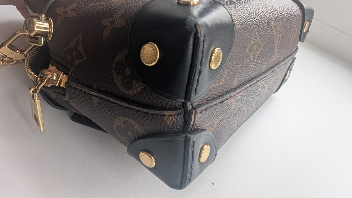 A close-up of a Louis Vuitton monogram canvas handbag corner with black leather trim and gold-tone hardware on a white surface. Pre-Owned Louis Vuitton Petite Malle Souple Monogram Bag