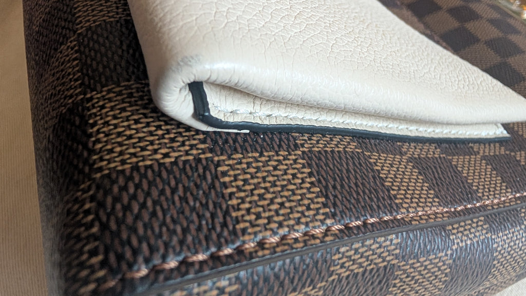 Close-up of a folded off-white textured leather item, likely a wallet or notebook cover, resting on a brown and beige checkered patterned surface. Pre-Owned Louis Vuitton Vavin PM Damier Ebene / Crème – N40113