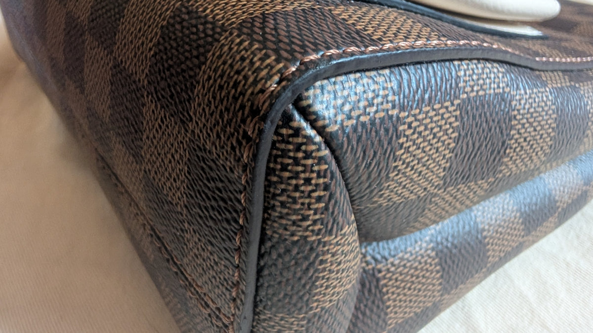 Close-up of a Louis Vuitton handbag corner showing the classic Damier Ebene checkered canvas in brown and beige with black leather trim. 