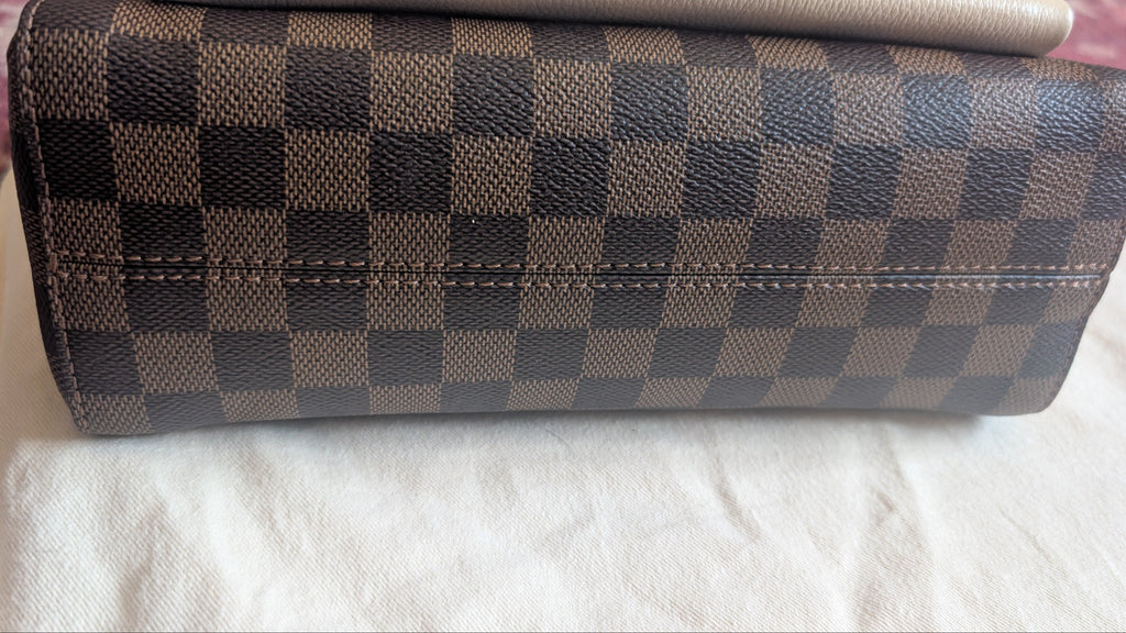 A Louis Vuitton clutch bag in the Damier Ebene checkered canvas, featuring a brown and beige checkerboard pattern, resting on a white fabric surface. Pre-Owned Louis Vuitton Vavin PM Damier Ebene / Crème – N40113