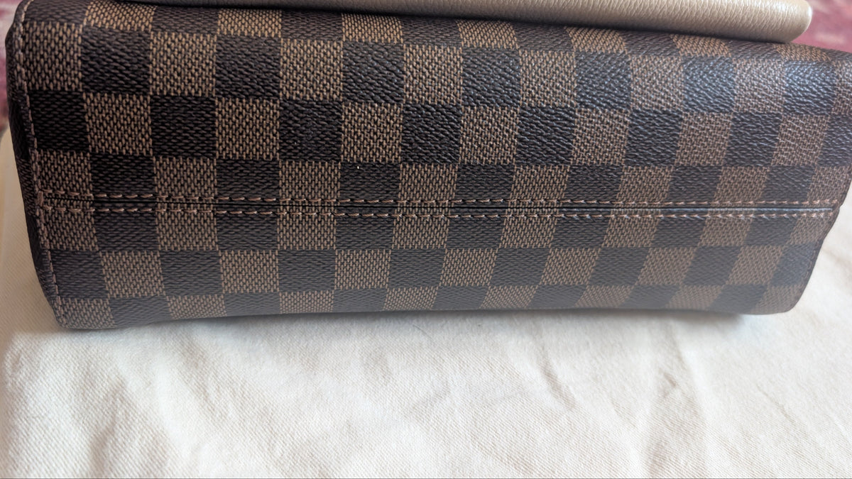 A Louis Vuitton clutch bag in the Damier Ebene checkered canvas, featuring a brown and beige checkerboard pattern, resting on a white fabric surface. Pre-Owned Louis Vuitton Vavin PM Damier Ebene / Crème – N40113