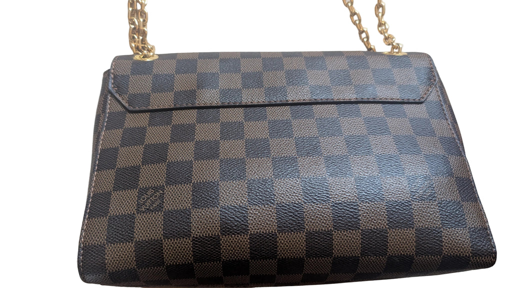A Louis Vuitton shoulder bag in Damier Ebene canvas, featuring a brown and gray checkerboard pattern with a gold-tone chain strap, laid flat on a light beige fabric background. Pre-Owned Louis Vuitton Vavin PM Damier Ebene / Crème – N40113