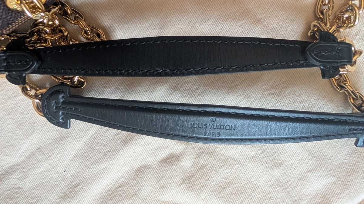 Close-up of a Louis Vuitton handbag strap featuring black leather trim and gold-tone chain links, laid flat on a beige fabric background. The Louis Vuitton brand name is embossed on the lower strap. Pre-Owned Louis Vuitton Vavin PM Damier Ebene / Crème – N40113