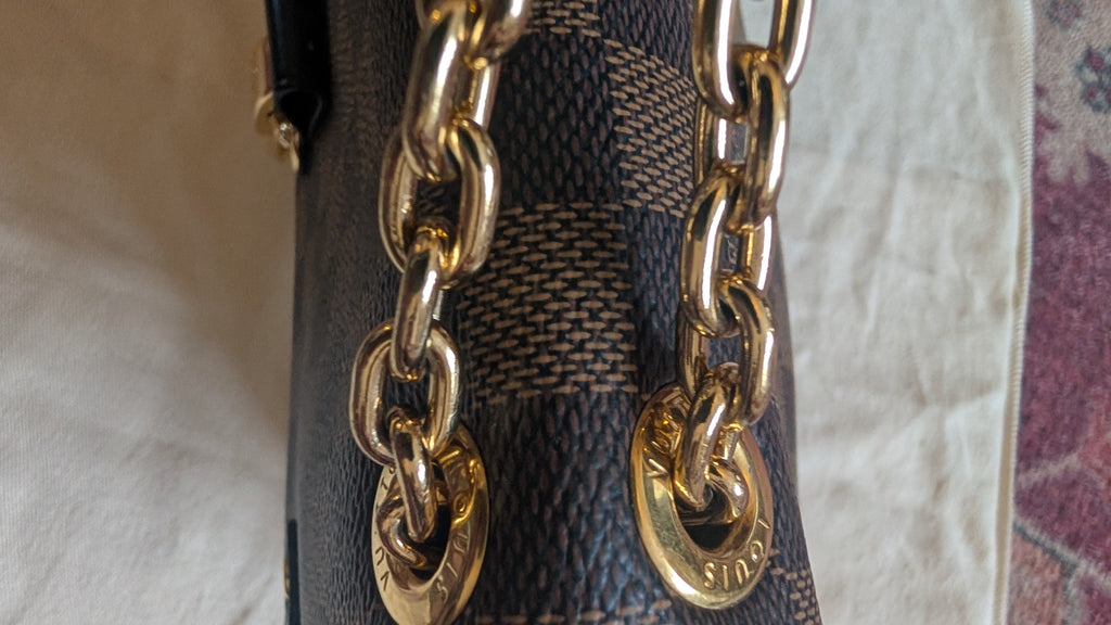 Close-up of a Louis Vuitton handbag featuring the brand's signature dark brown and beige checkered canvas, detailed with a prominent gold-tone chain strap and a gold-tone circular hardware charm engraved with 'LOUIS VUITTON'. Pre-Owned Louis Vuitton Vavin PM Damier Ebene / Crème – N40113