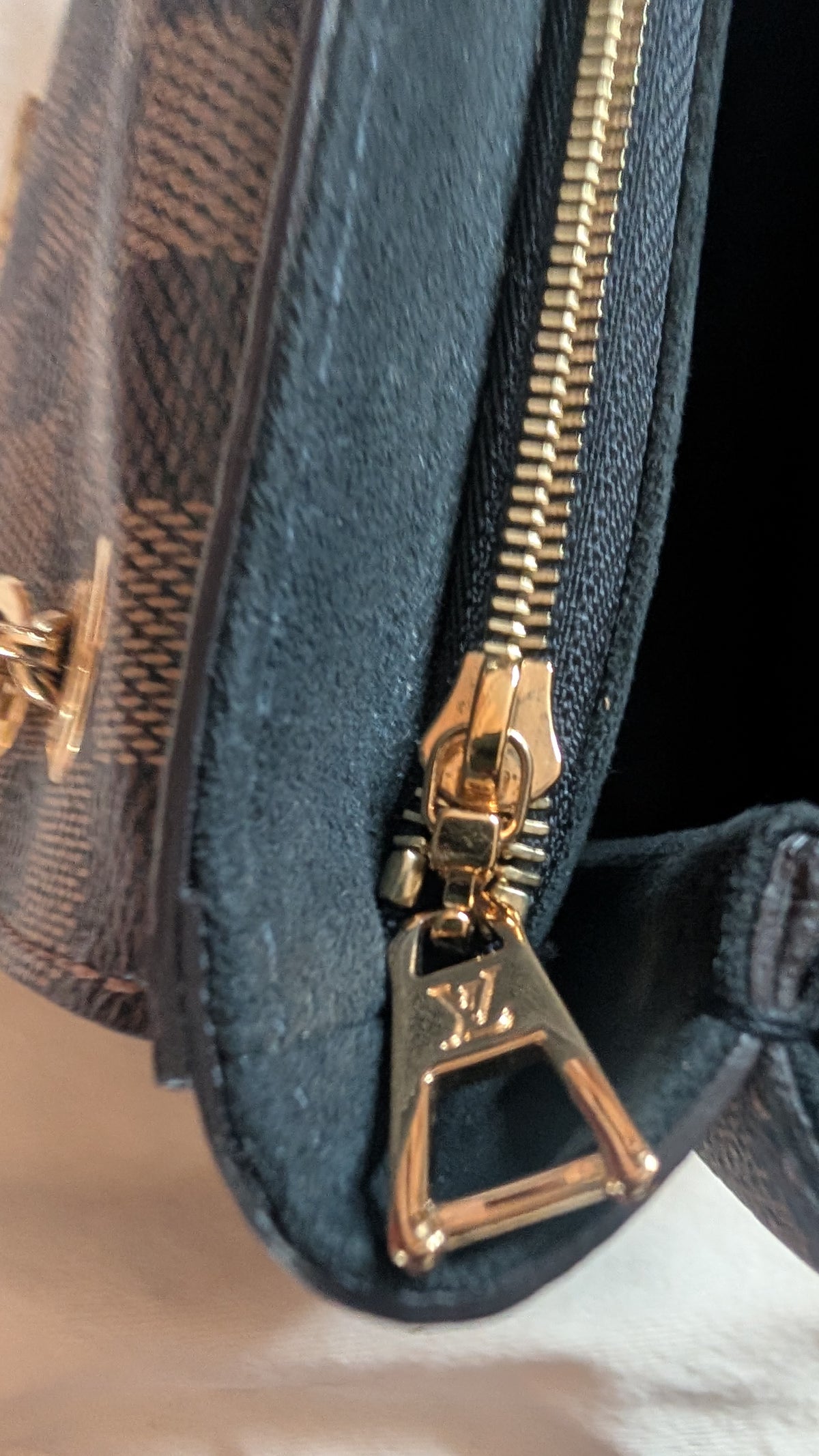 Close-up of a Louis Vuitton handbag corner showing dark brown monogram canvas, dark blue fabric trim, and a gold-tone zipper pull with the LV logo. Pre-Owned Louis Vuitton Vavin PM Damier Ebene / Crème – N40113